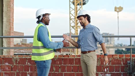 Multiethnic-men-in-hardhats-standing-at-the-construcing-site,-talking-and-shaking-hands.-Builder-and-foreman-meeting.-Outdoor.